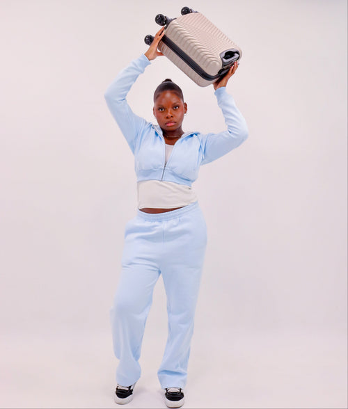 A model wearing a light blue zip-up hoodie and matching sweatpants holds a beige suitcase overhead against a plain light gray studio background.