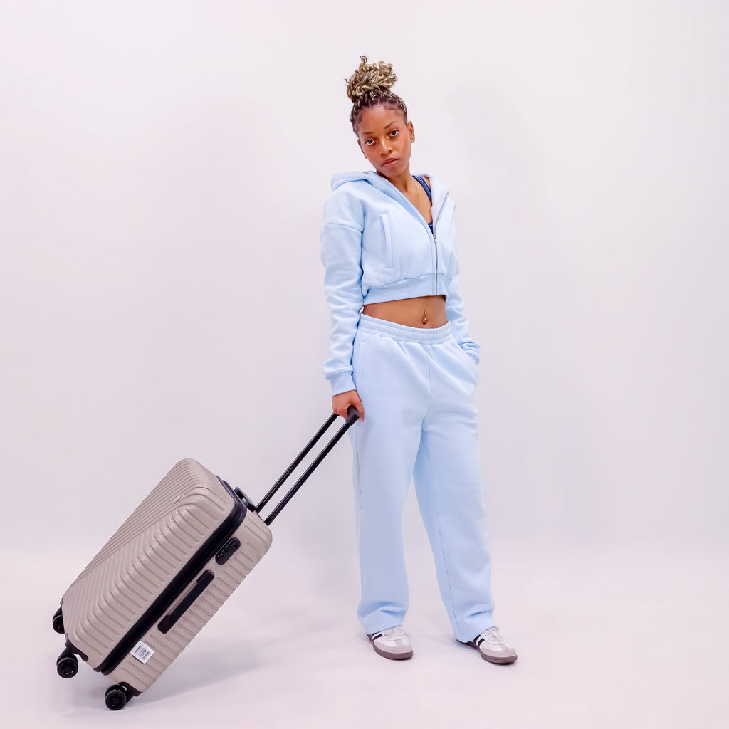 A woman posing in a light blue matching two-piece tracksuit, holding the handle of a silver hard-shell suitcase against a plain white studio background.